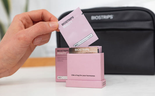 A person's hand holding a BIOSTRIPS™ Beauty pink packet labeled 'B01 Hormone Health' above a black pouch and several other pink packets. The background includes a white countertop and wooden object. Text overlay reads 'SUPPORTING YOUR JOURNEY TOWARDS HORM