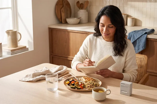 Adult woman recording food and digestive patterns beside water, ginger tea, a balanced meal and BIOSTRIPS® Glucose Support in a bright kitchen.