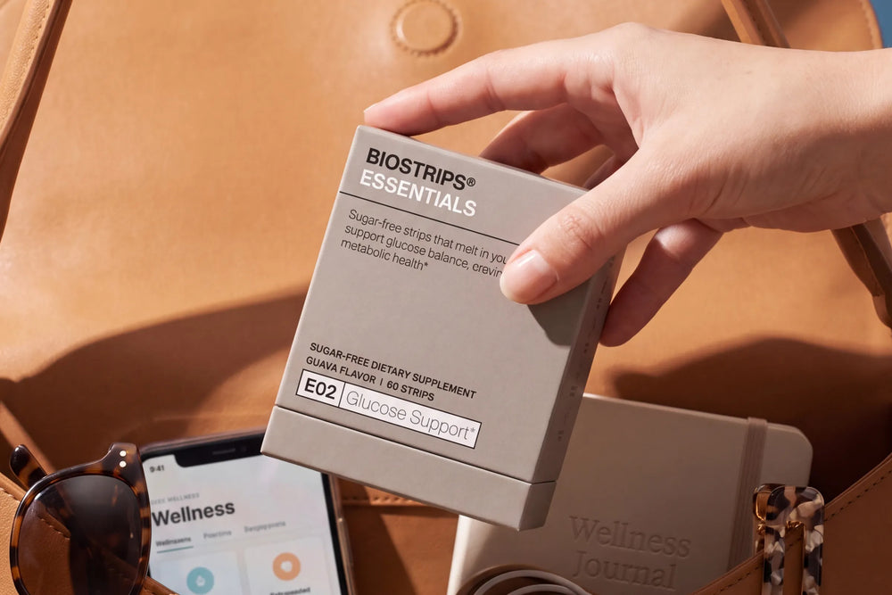 Hand holding a BIOSTRIPS Essentials Glucose Support carton over a tan bag with sunglasses and journal