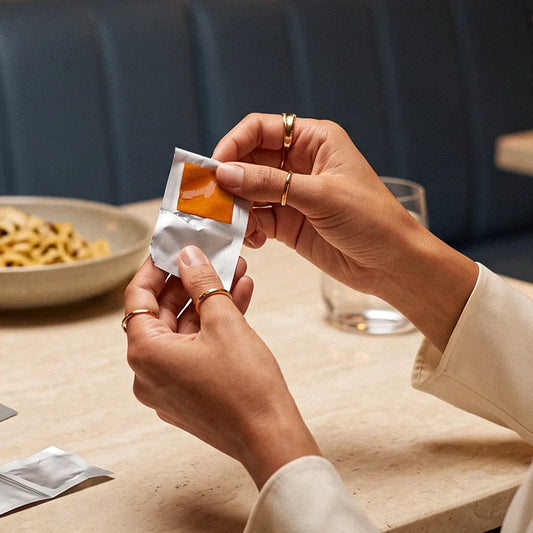 Close-up of a woman's hands opening a single-serve foil packet at a restaurant table, an orange strip visible inside